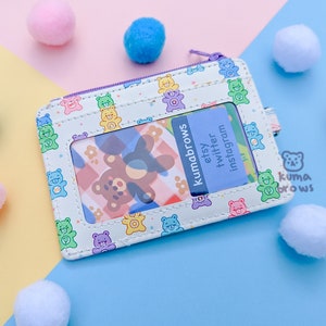 May include: A white cardholder with a purple zipper and a repeating pattern of colorful teddy bears. The cardholder has a clear window and a blue card with the text "kumabrows etsy twitter instagram". Pink, blue, and white pom-poms are scattered around.
