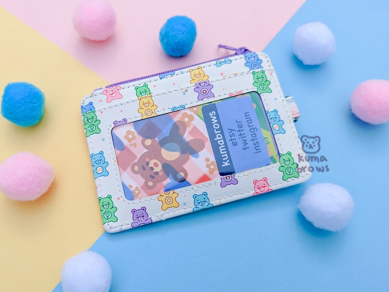 May include: A white cardholder with a purple zipper and a repeating pattern of colorful teddy bears. The cardholder has a clear window and a blue card with the text "kumabrows etsy twitter instagram". Pink, blue, and white pom-poms are scattered around.