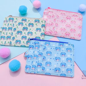 May include: Three small, rectangular wallets with zipper closures and multiple card slots. Each wallet features a repeating pattern of a cartoon character's face. The wallets are in light green, pink, and light blue, with matching pom-poms scattered around.