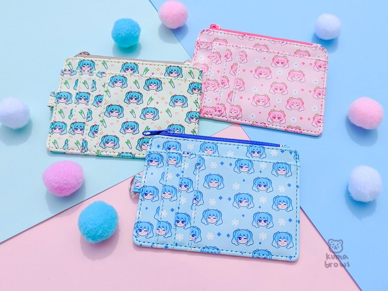 May include: Three small, rectangular wallets with zipper closures and multiple card slots. Each wallet features a repeating pattern of a cartoon character's face. The wallets are in light green, pink, and light blue, with matching pom-poms scattered around.