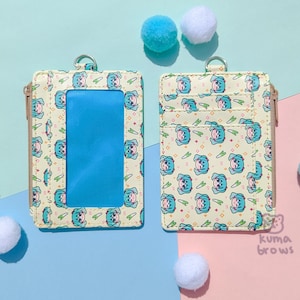 May include: Two cream-colored cardholders with a repeating cartoon character pattern and a clear ID window. Each cardholder has a zipper closure and a metal ring. The background is a pastel color scheme with fluffy pom-poms.