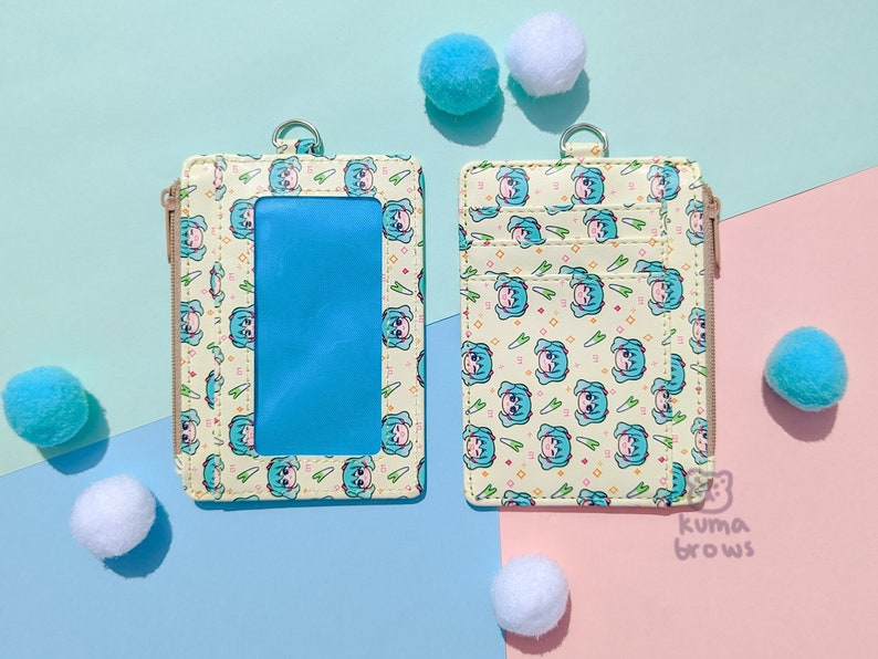 May include: Two cream-colored cardholders with a repeating cartoon character pattern and a clear ID window. Each cardholder has a zipper closure and a metal ring. The background is a pastel color scheme with fluffy pom-poms.