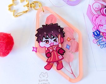 May include: A pink and orange diamond-shaped keychain with a cartoon character wearing a red jacket and a black headband. The keychain has dried pink flowers inside. The text "Kuma Brows" is printed on the bottom of the keychain.