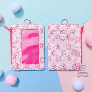 May include: Two pink cardholders with a repeating pattern of cartoon bears and flowers. One has a clear window and a pink zipper. The other has multiple card slots. Both have a metal ring at the top. Pink and white pom-poms are scattered around.