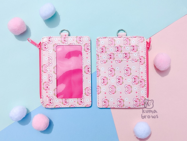May include: Two pink cardholders with a repeating pattern of cartoon bears and flowers. One has a clear window and a pink zipper. The other has multiple card slots. Both have a metal ring at the top. Pink and white pom-poms are scattered around.