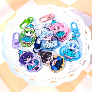 May include: Seven colorful character charms with keychains. Each charm features a different character wearing a hat and a green outfit. The charms are on a white plate with a small bear logo that says "Kuma Brows".