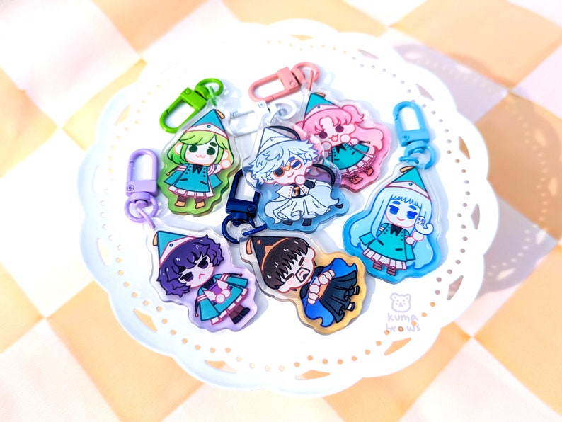 May include: Seven colorful character charms with keychains. Each charm features a different character wearing a hat and a green outfit. The charms are on a white plate with a small bear logo that says "Kuma Brows".