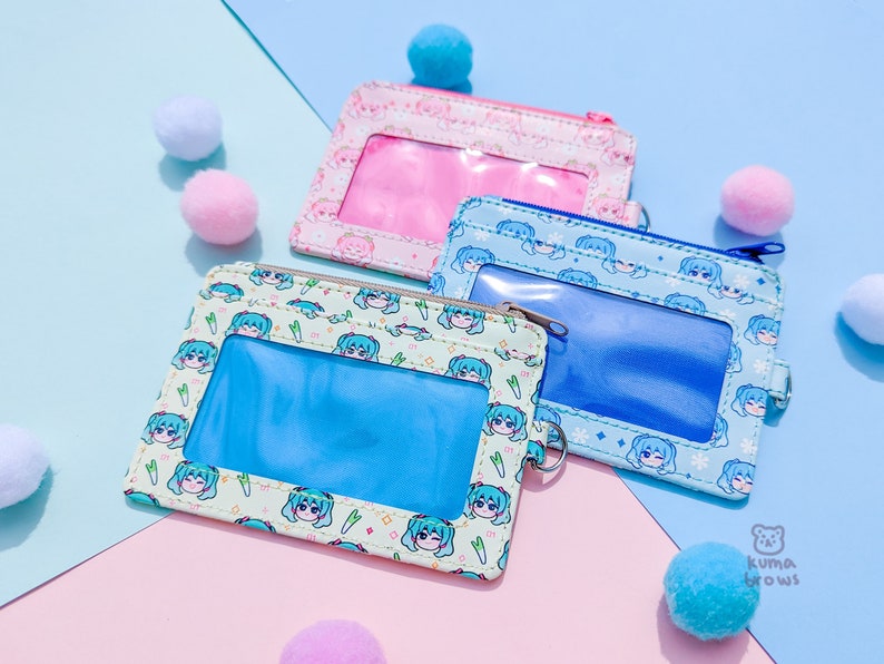 May include: Three colorful ID card holders with clear windows and zipper closures. The holders feature repeating patterns of anime-style characters in pink, blue, and mint green. Small pom-poms in matching colors are scattered around the holders.