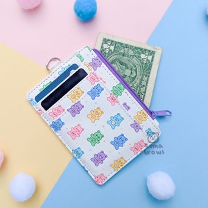 May include: White cardholder with a colorful bear pattern, a purple zipper, and a silver ring. The cardholder contains cards and a one-dollar bill. The background is a mix of pastel colors and decorative pom-poms.