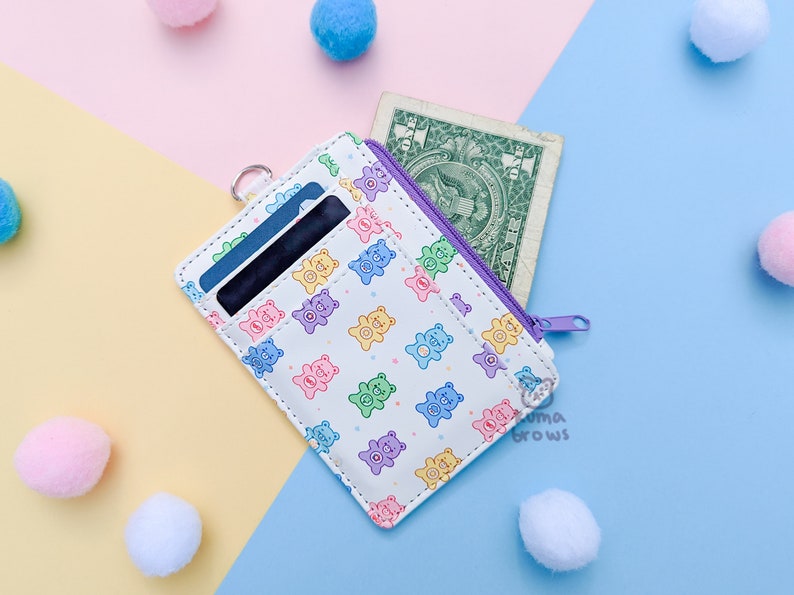 May include: White cardholder with a colorful bear pattern, a purple zipper, and a silver ring. The cardholder contains cards and a one-dollar bill. The background is a mix of pastel colors and decorative pom-poms.