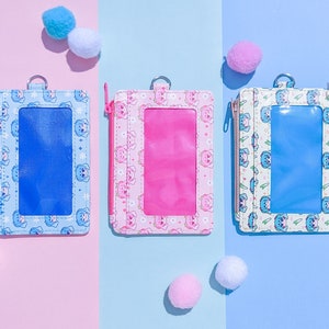 May include: Three rectangular ID holders in blue, pink, and white, each with a clear window and a zipper closure. The holders feature a repeating pattern of a cartoon bear and snowflake design. Small, fluffy pom-poms are scattered around the holders.