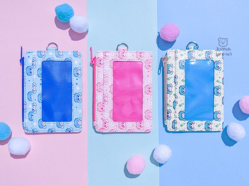 May include: Three rectangular ID holders in blue, pink, and white, each with a clear window and a zipper closure. The holders feature a repeating pattern of a cartoon bear and snowflake design. Small, fluffy pom-poms are scattered around the holders.