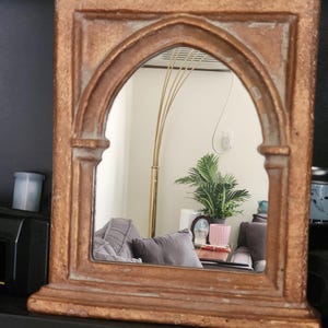May include: A rectangular, antique-style mirror with a gold-colored, ornate frame. The mirror has an arched top and reflects a room with a plant, lamp, and gray pillows. The frame has a textured, aged appearance.