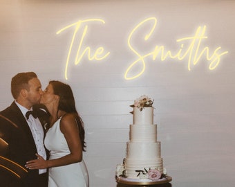 May include: A couple kisses in front of a white wall with a neon sign that reads "The Smiths". A white wedding cake with pink flowers sits on a gold cake stand on a black table.