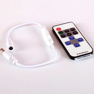 May include: A white remote control with a black face and buttons for controlling LED lights. The remote has buttons for speed, mode, and brightness. The remote also has a button for turning the lights on and off. The remote is connected to a white wire with a power adapter.