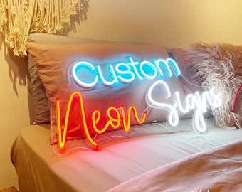 May include: A neon sign with the words "Custom Neon Signs" in blue, red, and white. The sign is on a clear backing and is placed on a bed with a pink comforter. The sign is lit up and the text is easy to read.