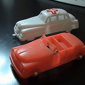 Puede incluir: Dos coches de juguete vintage, uno blanco con una cruz roja en el techo y otro rosa, ambos con detalles elaborados.