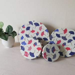 Puede incluir: Cuatro cubiertas de alimentos reutilizables de silicona con fondo blanco y un patrón de hojas de ginkgo azules y rosas. Las cubiertas son de diferentes tamaños.