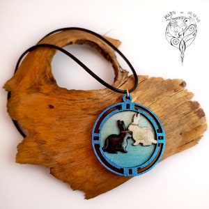 Könnte beinhalten: Eine blaue runde Anhänger-Halskette mit einem schwarzen und weißen Kaninchen-Design. Der Anhänger ist von einem dekorativen Rand umgeben. Die Halskette liegt auf einer Holzoberfläche.