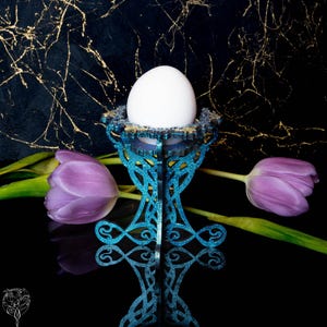 Könnte beinhalten: Ein weißes Ei liegt in einem teal-farbenen, glitzernden dekorativen Eierhalter mit einem verzierten Design. Zwei lila Tulpen mit grünen Stielen befinden sich auf beiden Seiten. Der Hintergrund ist schwarz mit goldenen Akzenten.
