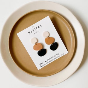 May include: A pair of dangle earrings featuring three geometric shapes in black, brown, and cream colours. The earrings are displayed on a white card with the text "The Masters Collective" and their social media handles.