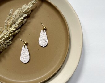 May include: A pair of white teardrop-shaped earrings with a textured floral design and gold accents. The earrings are displayed on a brown plate.