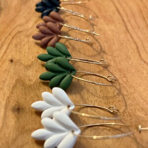 May include: Four pairs of gold hoop earrings with different colored clay flower charms. The charms are in shades of black, brown, green, and white.