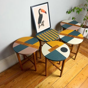 May include: Five small, round side tables with wooden legs and geometric patterns in teal, mustard yellow, pink, and wood tones. A framed toucan print leans against one table. The tables are ideal for a living room or bedroom setting.