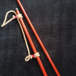 Könnte beinhalten: Zwei rote Holzstangen mit einem braunen Seil, das um sie herumgebunden ist. Das Seil hat rote Knoten.