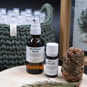 Könnte beinhalten: Eine braune Glassprayflasche und eine kleine Ölflasche mit weißen Etiketten, beschriftet mit "Bio Zirben Spray" und "Bio Zirben Öl". Ein Tannenzapfen und Kiefernnadelzweige liegen auf einer Holzoberfläche. Ein grüner Korb enthält weitere Sprühflaschen.