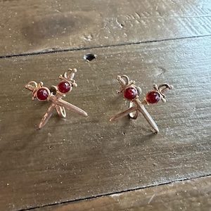 May include: A pair of gold-toned metal clip-on earrings shaped like two crossed swords. Each sword hilt is accented with a small, round, faceted red gemstone. The earrings feature a metallic finish and a classic sword design with detailed hilts.