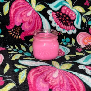 Puede incluir: Una vela rosa en un tarro Ball. El tarro de cristal transparente tiene el logotipo de Ball en relieve en la parte delantera. La vela es de color rosa sólido. El fondo es una tela negra con un estampado floral en rosa, turquesa y amarillo.