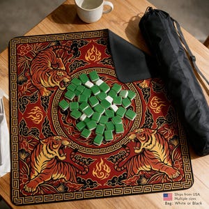 May include: A square mahjong mat featuring a red background with a gold geometric border and three illustrated tigers surrounded by stylized clouds and flames. A pile of green and white mahjong tiles sits in the center, accompanied by a black carrying bag.
