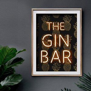 May include: A framed print with a gold border, featuring the words "THE GIN BAR" in a neon-style font. The background is black with a repeating gold pineapple pattern. The print is mounted on a dark gray wall.