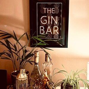May include: A framed sign with the words "THE GIN BAR" in white neon against a black background with gold pineapple illustrations. The sign hangs above a bar cart with cocktail glasses, a shaker, and bottles of liquor.