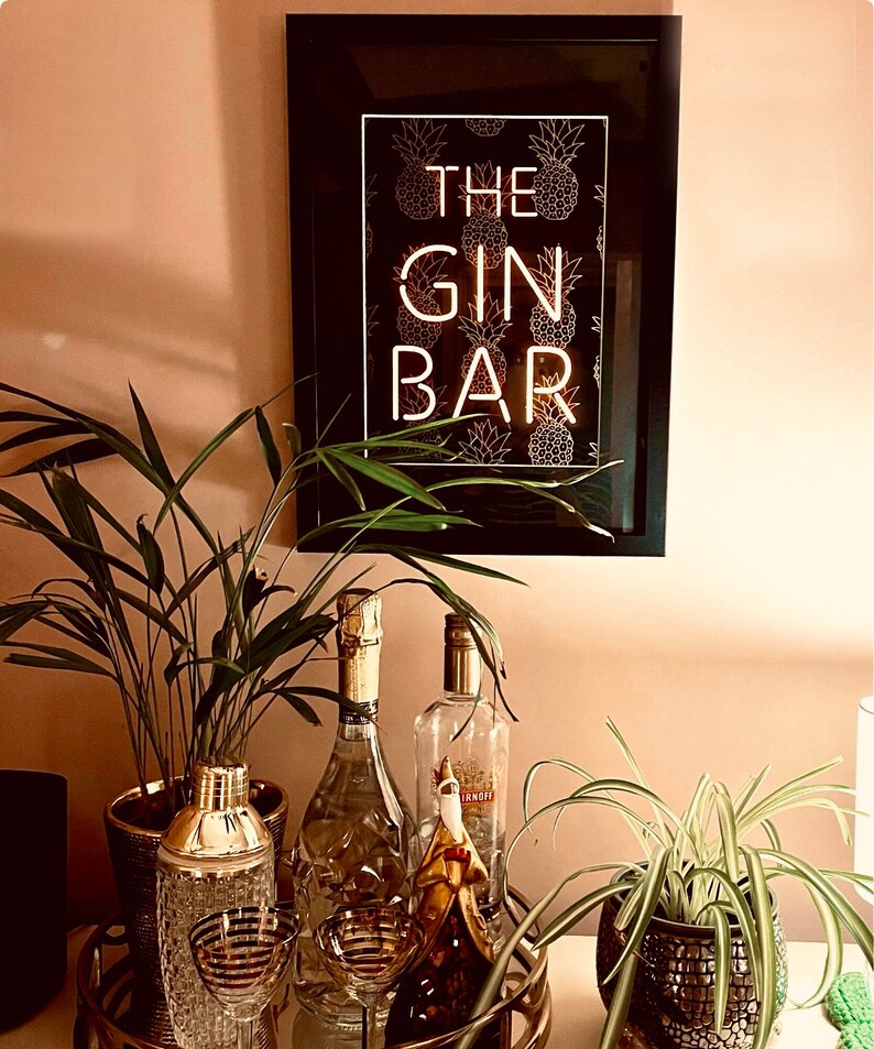 May include: A framed sign with the words "THE GIN BAR" in white neon against a black background with gold pineapple illustrations. The sign hangs above a bar cart with cocktail glasses, a shaker, and bottles of liquor.