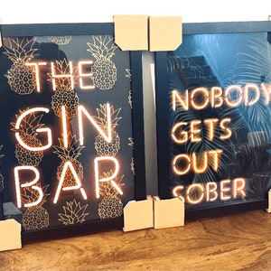 May include: Two framed art prints with black frames. One features the text "THE GIN BAR" in neon-style lettering over a pineapple pattern. The other says "NOBODY GETS OUT SOBER" in neon lettering over a tropical leaf design.