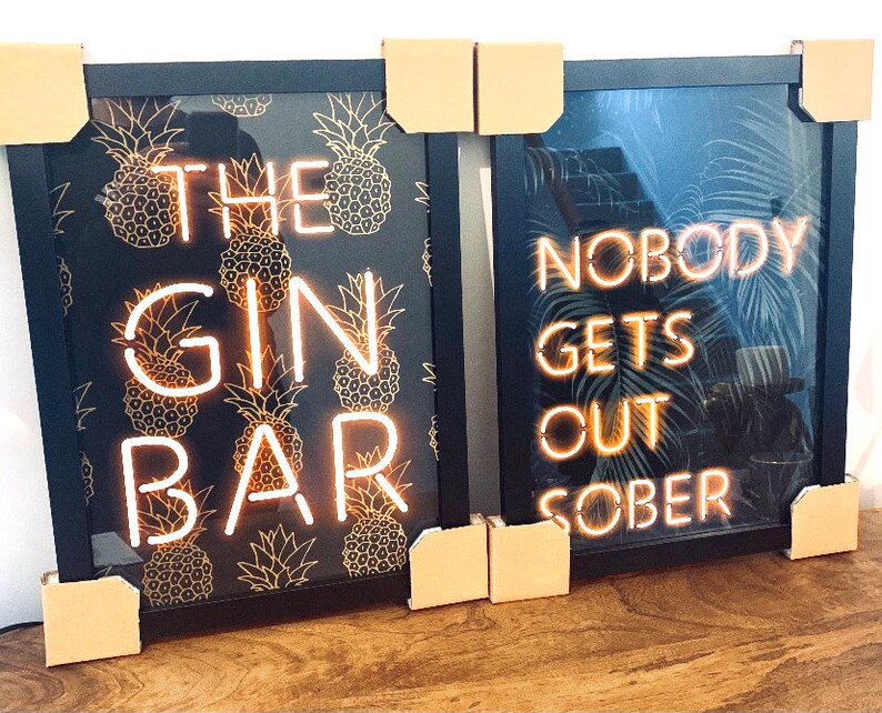May include: Two framed art prints with black frames. One features the text "THE GIN BAR" in neon-style lettering over a pineapple pattern. The other says "NOBODY GETS OUT SOBER" in neon lettering over a tropical leaf design.