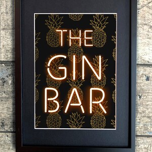 May include: Framed art print featuring the words "THE GIN BAR" in neon-style lettering. The background is black with a repeating pattern of gold pineapple illustrations. The frame is black.