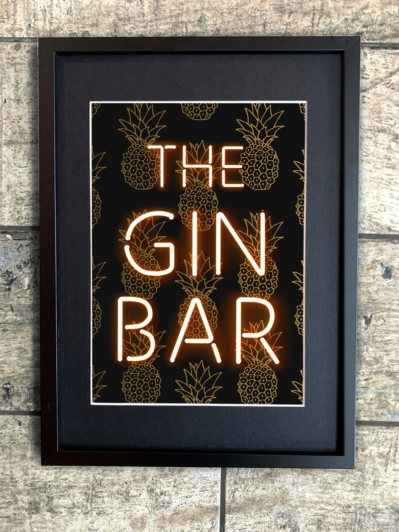 May include: Framed art print featuring the words "THE GIN BAR" in neon-style lettering. The background is black with a repeating pattern of gold pineapple illustrations. The frame is black.