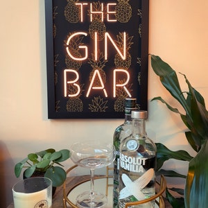 May include: A framed sign with the words "THE GIN BAR" in neon lights, set against a black background with gold pineapple patterns. A round gold tray holds a cocktail glass and a bottle of Absolut Vanilla vodka. A candle and a plant are also visible.