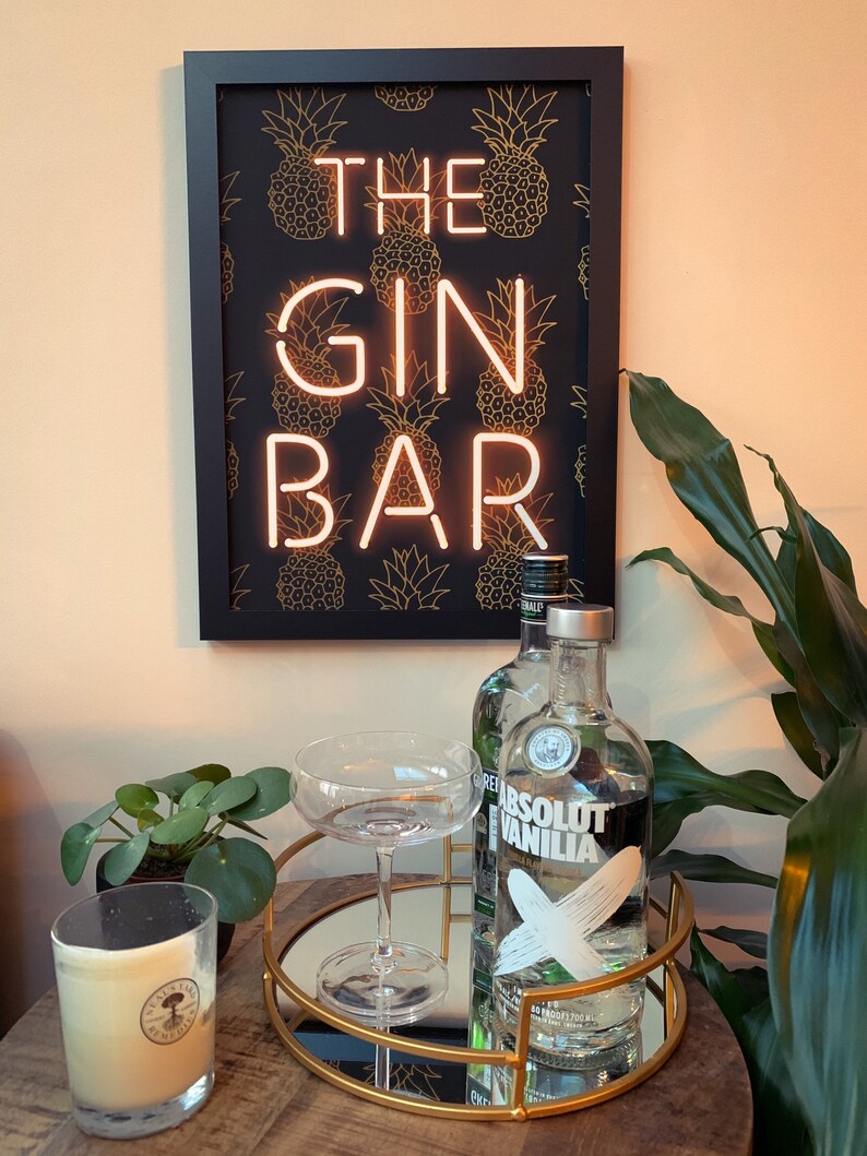 May include: A framed sign with the words "THE GIN BAR" in neon lights, set against a black background with gold pineapple patterns. A round gold tray holds a cocktail glass and a bottle of Absolut Vanilla vodka. A candle and a plant are also visible.