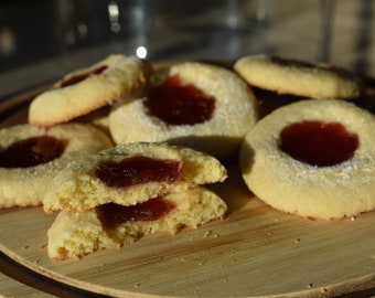 May include: A close-up of a plate of homemade jam-filled cookies. The cookies are round and have a light brown color. The jam filling is a bright red color. The cookies are dusted with powdered sugar.