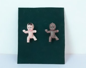 Op de afbeelding: Twee kleine plastic babyfiguurtjes, een met een lichte huid en de andere met een bruine huid, worden getoond op een donkergroene achtergrond. Beide figuurtjes dragen luiers en hebben hun armen uitgestrekt.