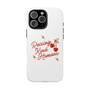 May include: White phone case with a red design that reads "Raising Kind Humans" in a cursive font. The design includes two cherries and star accents. The phone case has a black camera module.