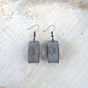May include: A pair of gray, rectangular earrings with a distressed finish and a geometric design in the center. The earrings have a hook closure and a rustic, vintage aesthetic. The earrings are displayed on a white surface.