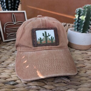 May include: A tan baseball cap featuring a rectangular leather patch on the front. The patch contains a cross-stitched design of green cacti against a light blue sky and orange desert landscape, framed by dark brown stitched leather edges.
