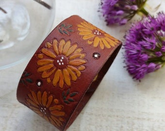 May include: A brown leather bracelet with a floral design featuring yellow and white flowers. The bracelet is on a white surface with purple flowers in the background.