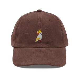 Könnte beinhalten: Braune Cord-Baseballkappe mit gesticktem Kakadu-Design. Der Kakadu hat einen gelben Kamm, einen weißen Körper und gelbe und rote Akzente. Die Kappe hat einen gebogenen Schirm und einen verstellbaren Stoffriemen.