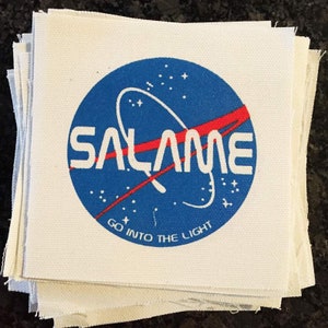 Puede incluir: Un parche de tela blanco con un círculo azul que contiene una línea curva roja y la palabra "SALAME" en letras blancas. El círculo también tiene estrellas blancas y el texto "GO INTO THE LIGHT" en letras blancas.